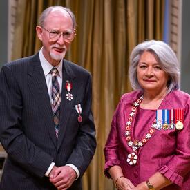 La gouverneure générale Simon se tient à côté d’un homme qui porte une médaille épinglée à son blazer gris.