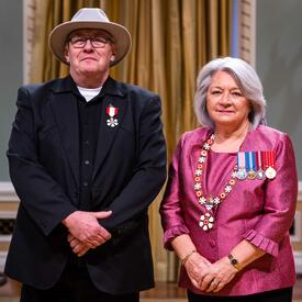 La gouverneure générale Simon se tient à côté d’un homme qui porte une médaille épinglée à son blazer noir. Il porte un chapeau.