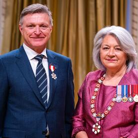 La gouverneure générale Simon se tient à côté d’un homme qui porte une médaille épinglée à son blazer bleu.