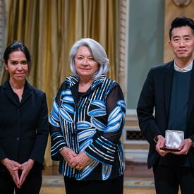 Governor General Simon is standing in between a man and a woman. The man is holding a small box with a medallion inside of it.
