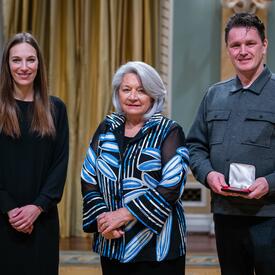 La gouverneure générale Simon se tient entre un homme et une femme. L'homme tient une petite boîte dans laquelle se trouve une médaille.