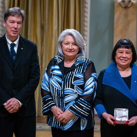 Governor General Simon is standing in between a man and a woman. The woman is holding a small box with a medallion inside of it.