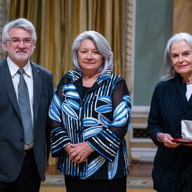 La gouverneure générale Simon se tient entre un homme et une femme. La femme tient une petite boîte dans laquelle se trouve une médaille.