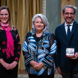La gouverneure générale Simon se tient entre un homme et une femme. L'homme tient une petite boîte dans laquelle se trouve une médaille.