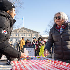 Governor General Simon speaks to a person standing behind a table. There is a colourful game on the table. They are outdoors and wearing winter clothing. 