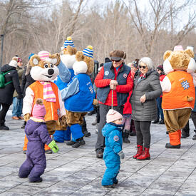 Governor General Simon and Mr. Fraser are smiling at a group of children. The Winterlude Ice Hogs and a group of people stand behind them.