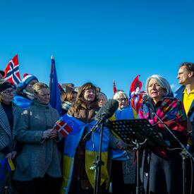Governor General Simon speaking into a microphone. A crowd is gathered around her. Many people are holding flags from different countries.