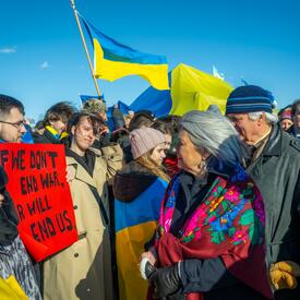 Governor General Simon and Mr. Fraser walk through a crowd of people. She looks at a man holding a red sign that reads, “If we don’t end war, war will end us.” Many other people are holding Ukrainian flags.
