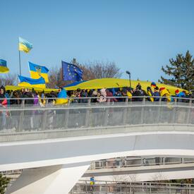 A group of people stand on a bridge. They are holding a large, yellow-and-blue Ukrainian flag above them. Others are waving smaller Ukrainian flags. A line of trees and blue sky are behind them.