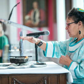 An Inuk Elder wearing traditional clothing holds her hand above a lit qulliq (oil lamp) as part of its inaugural lighting ceremony at Rideau Hall.  