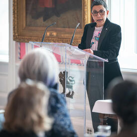 A woman stands at a podium and speaks into a microphone in the Tent Room at Rideau Hall. A group of women sit in the foreground, listening to her speak. 