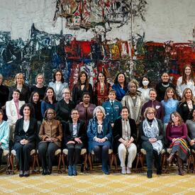 A group of approximately 40 women pose with Governor General Simon for a photo in the Ballroom at Rideau Hall. They are gathered in front of a large painting.