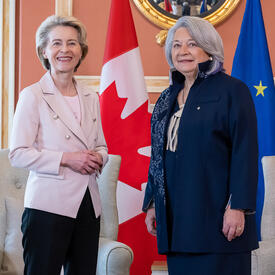 Governor General Simon Stands next to Her Excellency Ursula von der Leyen, President of the European Commission. They are smiling at the camera. Behind them is the flag of Canada and the flag of Europe.