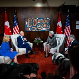 Governor General Simon, Mr. Whit Fraser, President Joe Biden, and First Lady Jill Biden seated in grey armchairs facing one another. Two Canada flags and two American flags are behind them. Several people with cameras are filming them.