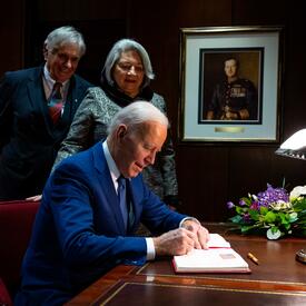 Le président Joe Biden signe un livre d'or. La gouverneure générale Simon et M. Whit Fraser se tiennent derrière lui.