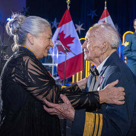 La gouverneure générale Mary Simon tient le général Richard Rohmer dans ses bras pendant que les membres de la Musique centrale des Forces canadiennes regardent en arrière-plan