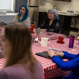 A group of people sit attentively around a table with a red-checkered tablecloth in a kitchen setting.