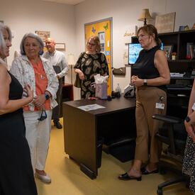  A group of five people converse in an office, surrounded by shelves and a desk.