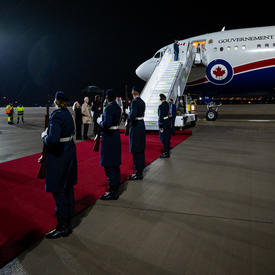 The Government of Canada plane is on the tarmac at the airport. The door to the plane is open and the stairs are open waiting for the passengers to descend. There is a red carpet at the base of the plane.