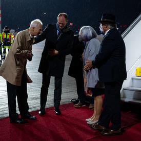 Governor General Mary Simon and Mr. Whit Grant Fraser are greeted by German and Canadian officials. They are on a red carpet at the foot of a plane. There are people in military uniform along the red carpet. It is nighttime. 