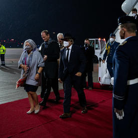 Governor General Mary Simon and Mr. Whit Grant Fraser are greeted by German and Canadian officials. They are on a red carpet at the foot of a plane. There are people in military uniform along the red carpet. It is nighttime. 