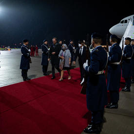Governor General Mary Simon and Mr. Whit Grant Fraser are greeted by German and Canadian officials. They are on a red carpet at the foot of a plane. There are people in military uniform along the red carpet. It is nighttime. 