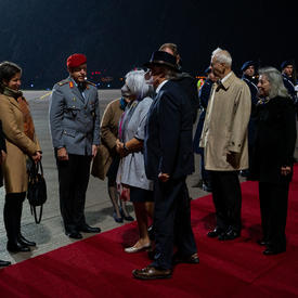Governor General Mary Simon and Mr. Whit Grant Fraser are greeted by German and Canadian officials. They are on a red carpet at the foot of a plane. There are people in military uniform along the red carpet. It is nighttime. 