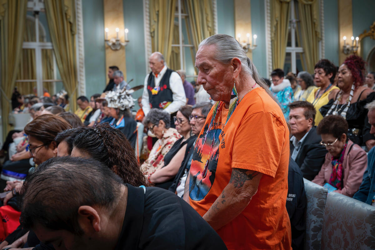 Un homme aux cheveux argentés vêtu d'une chemise orange se tient debout parmi des personnes assises