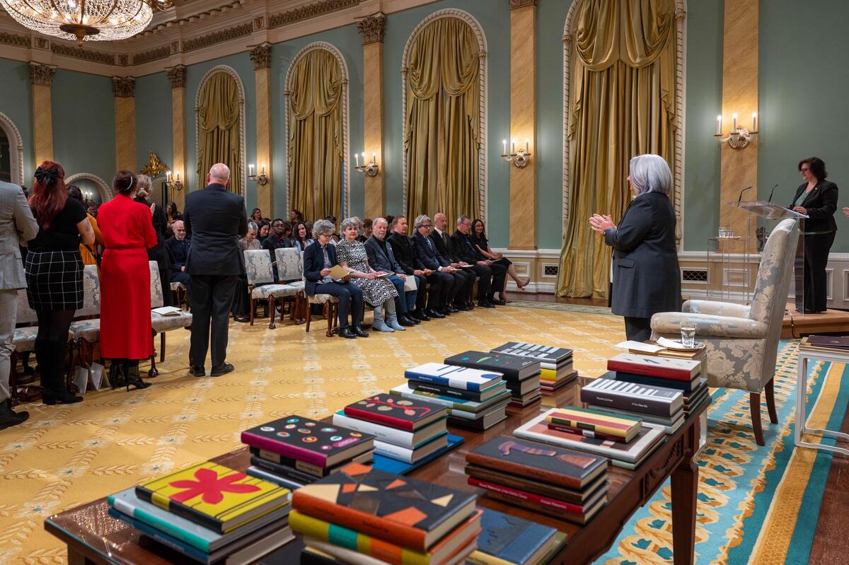 Governor General Mary Simon claps as recipients enter the Ballroom. In front their is a table whit books