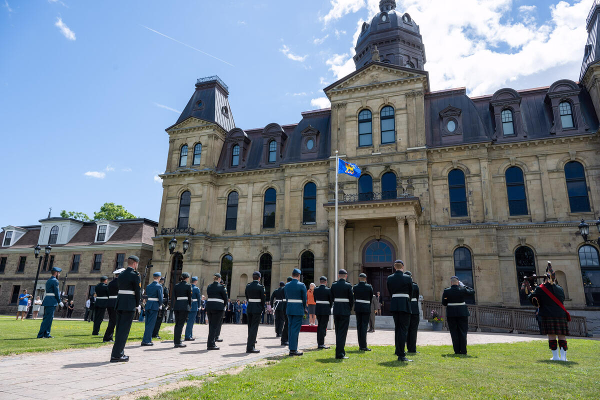 Un groupe de personnes en uniforme militaire se tient en rangs et fait face à un grand bâtiment en pierre. Le drapeau vice-royal flotte sur un mât de drapeau.