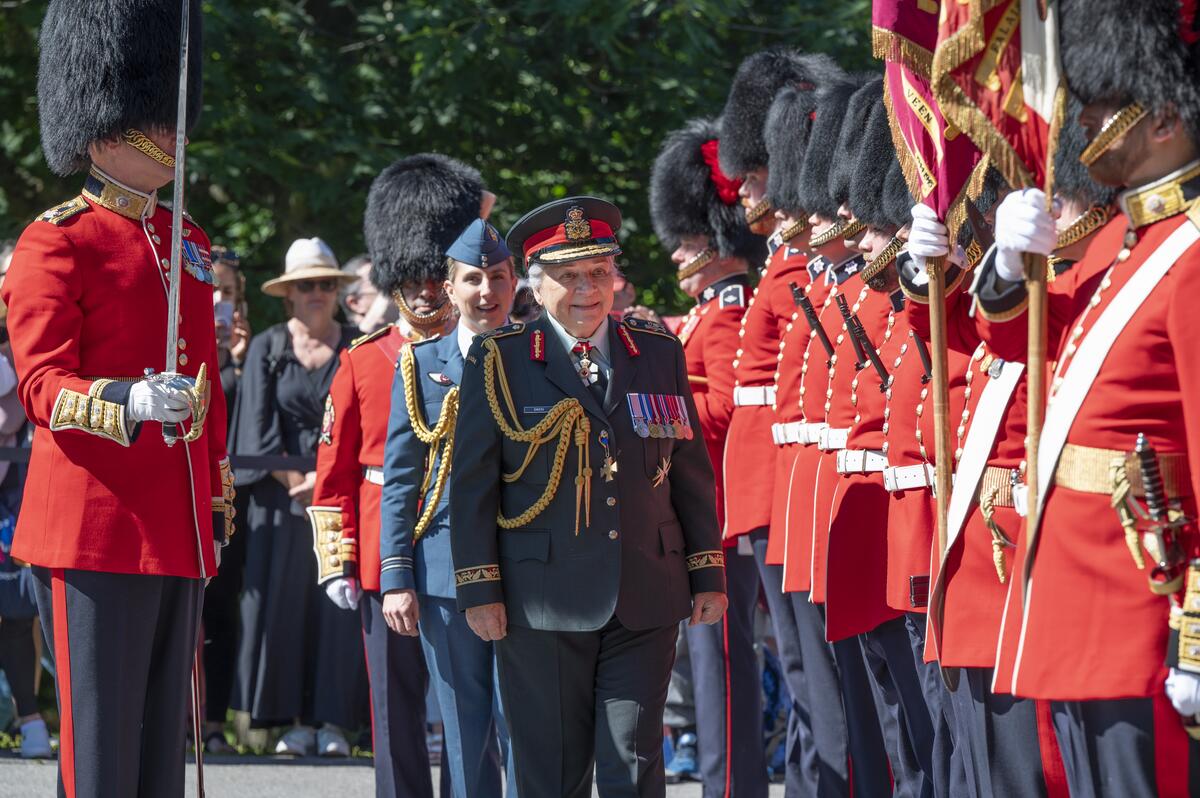 Governor General Mary Simon inspects the guard. A member of the Royal Canadian Air Force is behind her. She is also wearing a Canadian Army uniform. Members of the guard are to her left, all wearing red uniforms.