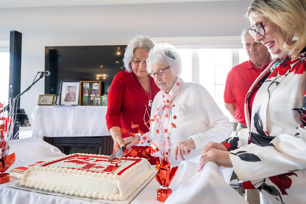 Des gens sont vêtus de rouge et de blanc pour célébrer la fête du Canada dans une maison de retraite. La gouverneure générale Simon aide une femme à couper un gros gâteau.