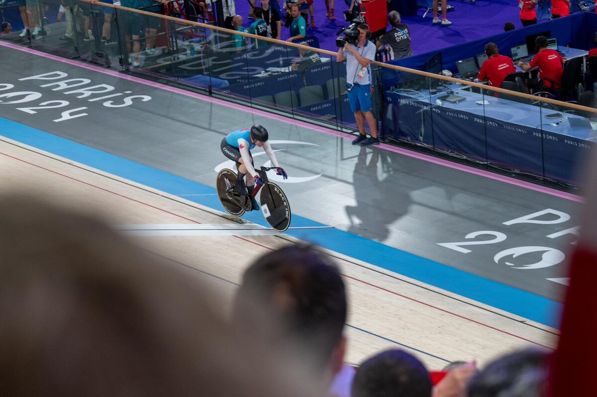 A Team Canada paracyclist competes in a track event at the Paris 2024 Paralympics. The athlete is on a track bicycle, wearing a blue uniform and helmet. The "Paris 2024" logo is visible on the floor.