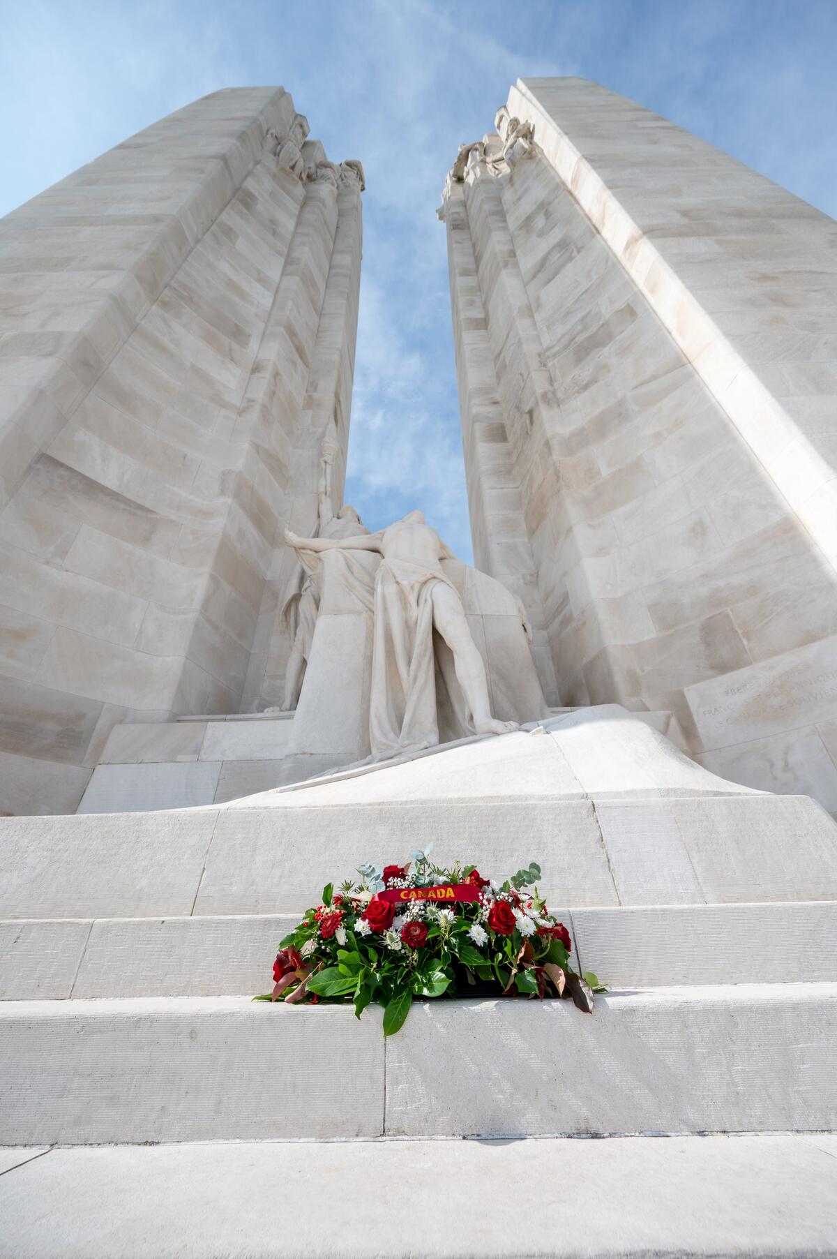 A wreath of red, white, and green flowers lies on the steps in front of a large, white stone monument with two tall pillars and a sculpted figure.