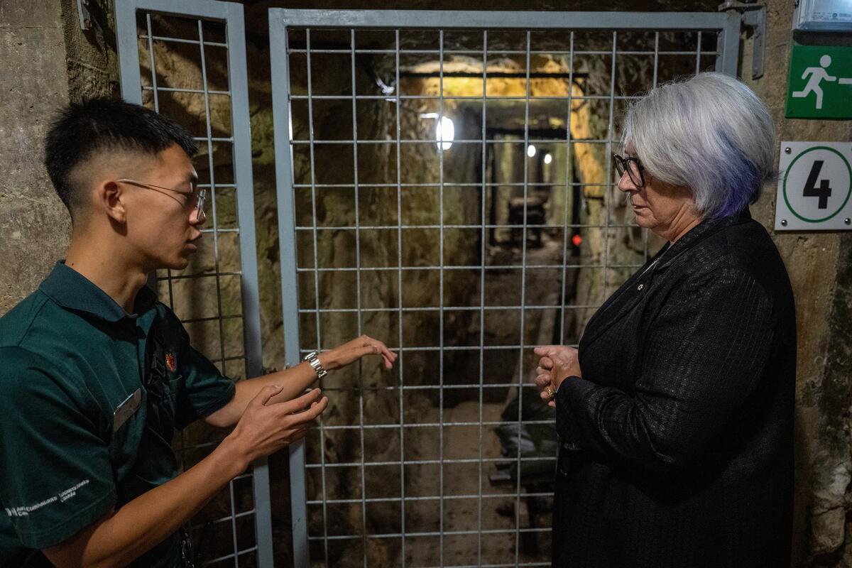 A person in a green shirt is speaking to the Governor General of Canada Mary Simon who is wearing a black jacket. They stand in front of a gated entrance to an underground stone corridor.