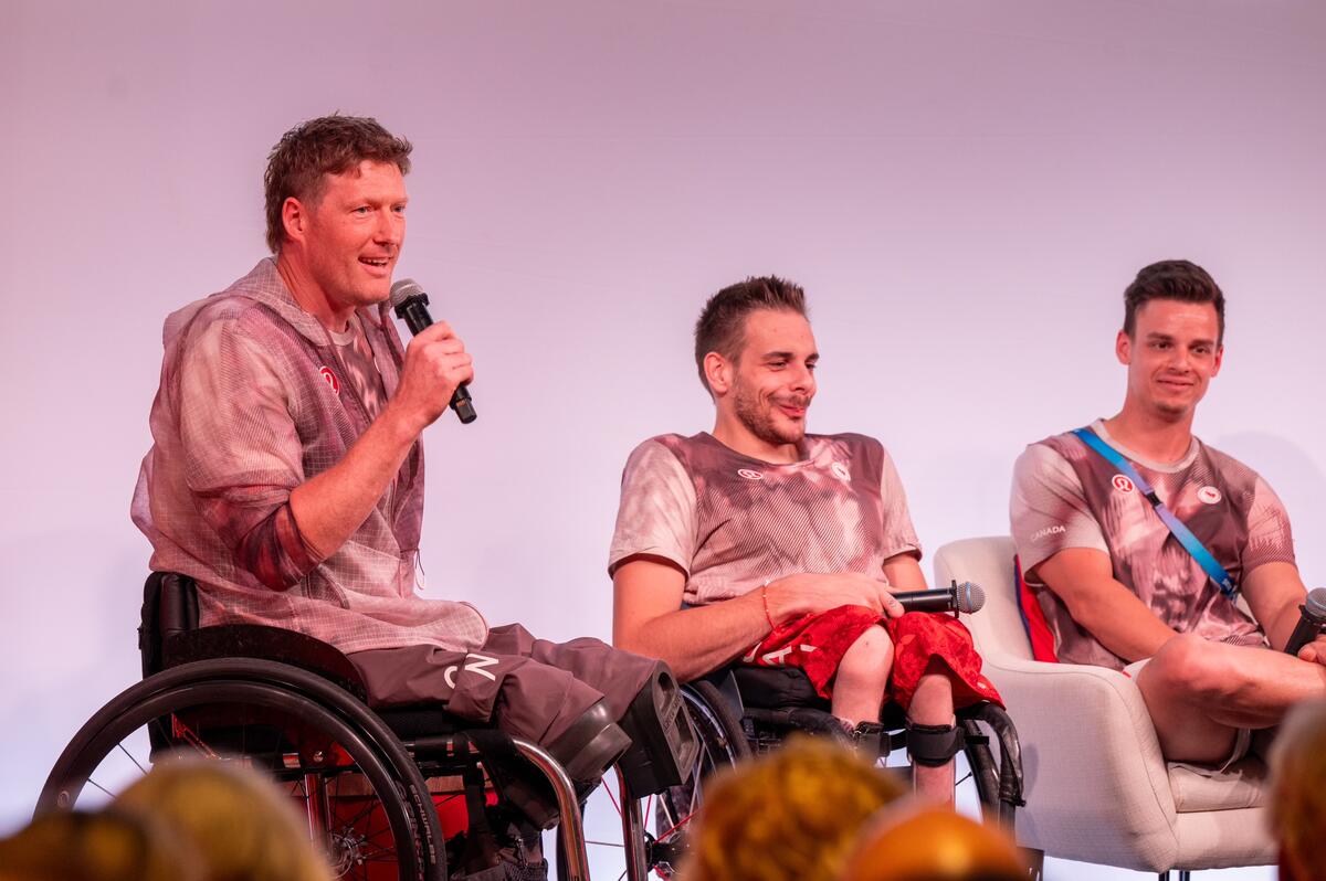 Three people, including two in wheelchairs, are participating in a discussion. The person on the left is speaking into a microphone. An audience is visible in the foreground.