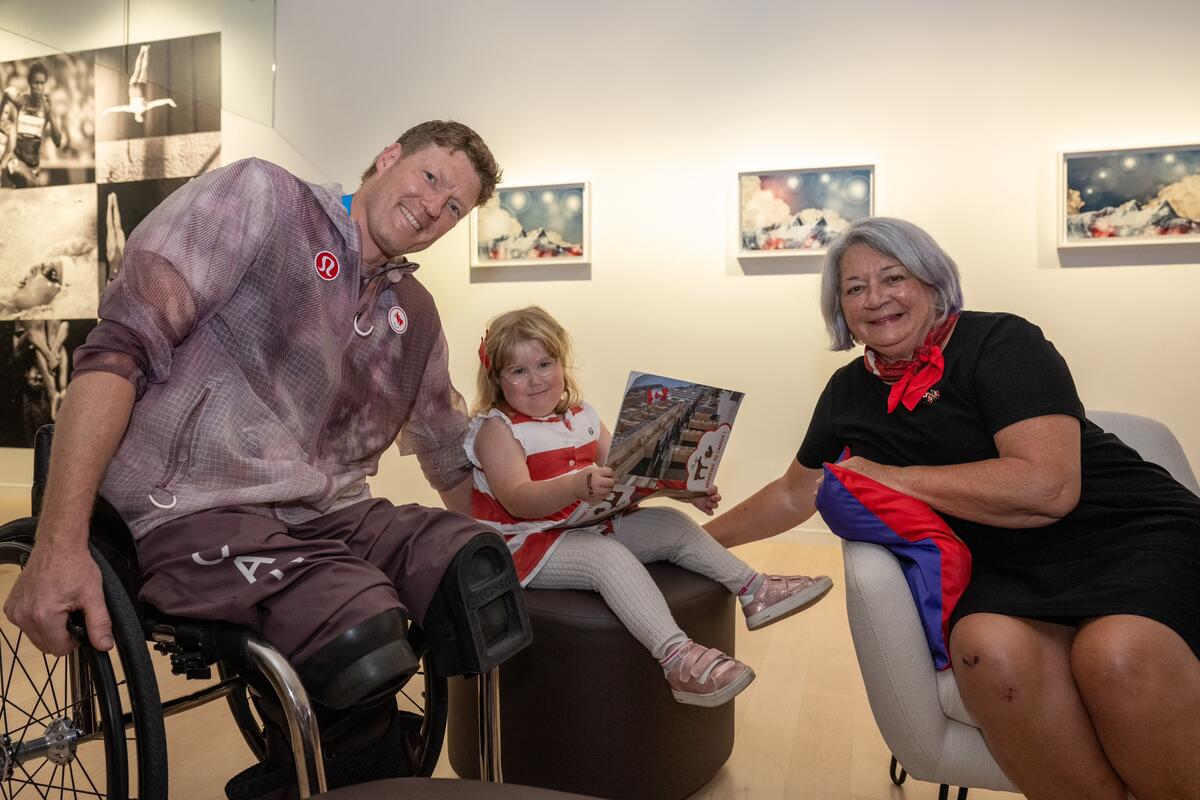 A person in a wheelchair and the Governor General of Canada Mary Simon sit indoors with a young girl seated between them, holding a book. All three are smiling. 