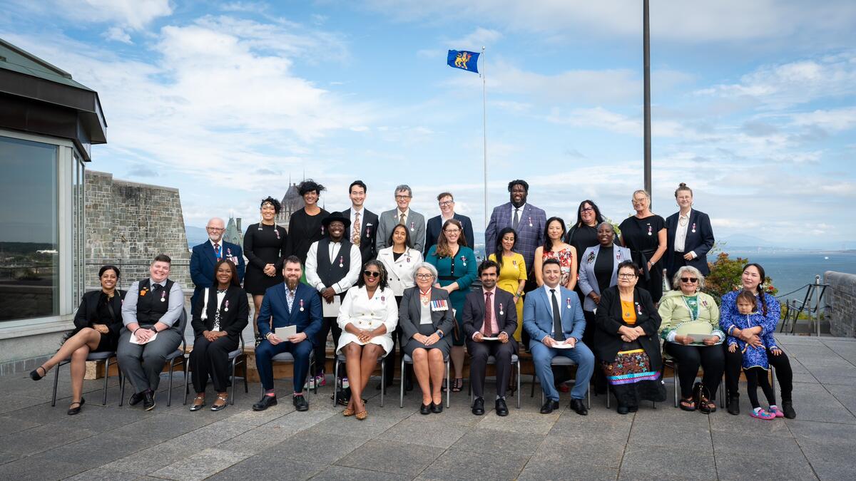 Un groupe de personnes portant des médailles assis sur des chaises à l'extérieur avec la gouverneure générale. Ils sont dehors.