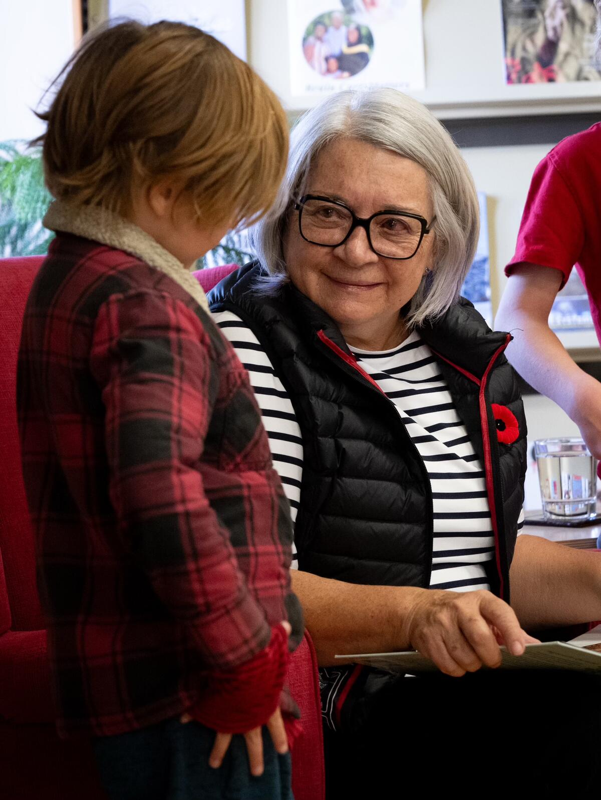 La gouverneure générale Mary Simon est assise sur une chaise rouge, elle tient un livre et sourit à un enfant qui se tient à ses côtés.