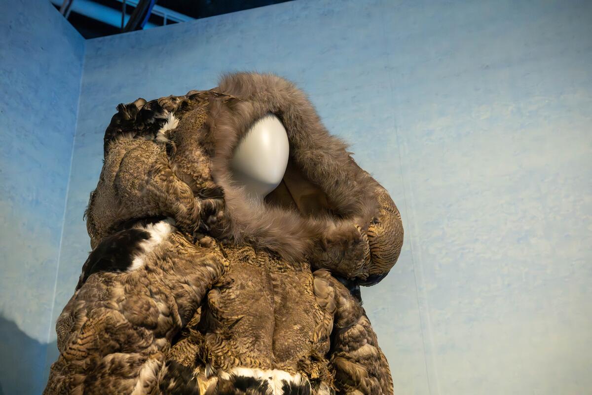 Mannequin vêtu d'une parka en fourrure brune texturée avec une grande capuche, sur fond bleu clair. La scène dégage une atmosphère froide et arctique.
