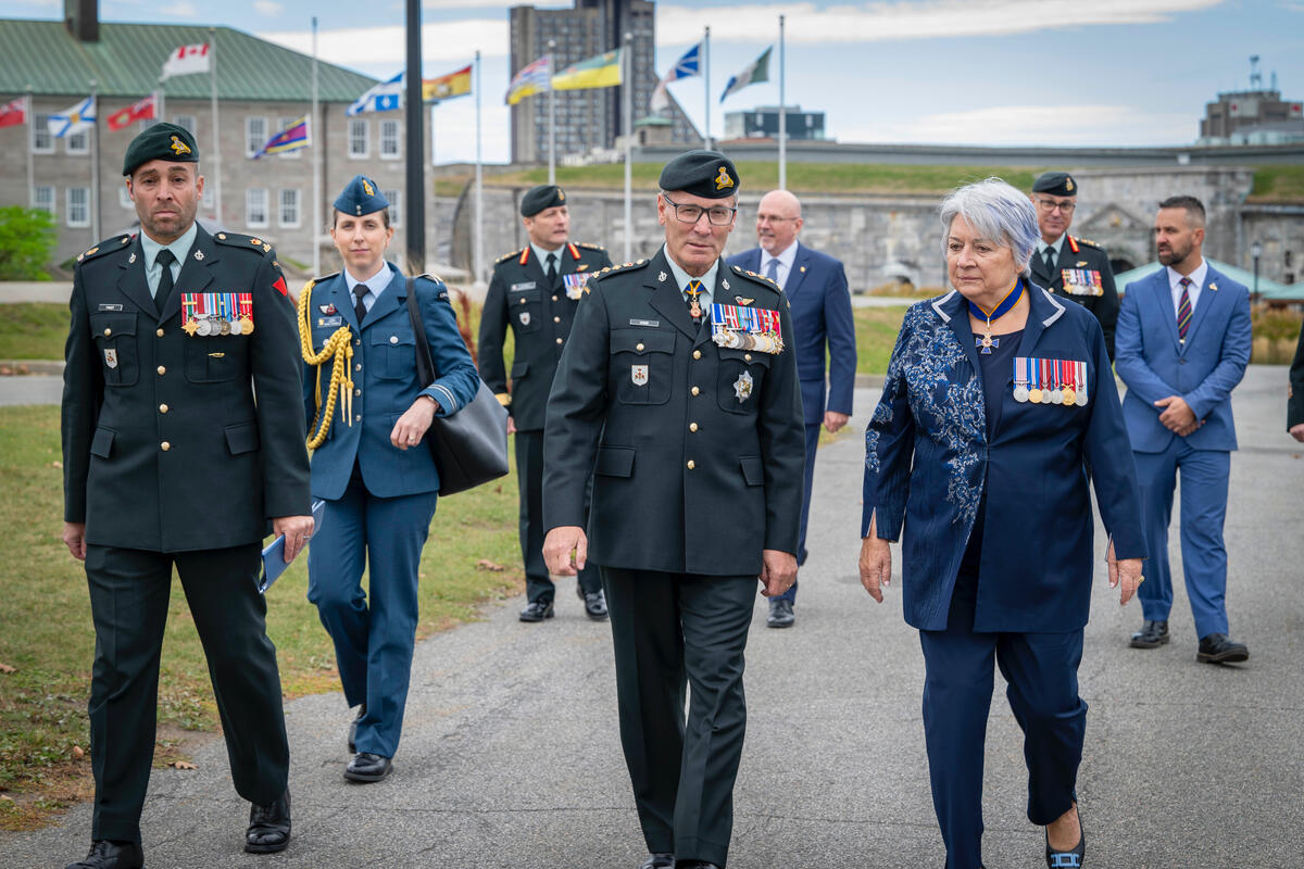 Governor General Mary Simon walking with a group of military personnel and officials, wearing uniforms with medals, on a path. Flags and historic buildings are visible in the background.
