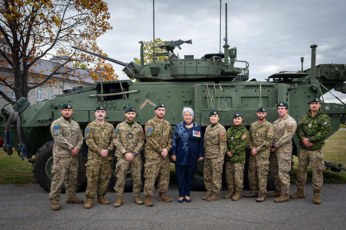 Governor General Mary Simon posing for a photo with a group of military personnel in camouflage uniforms and berets. A large army tank is behind them.