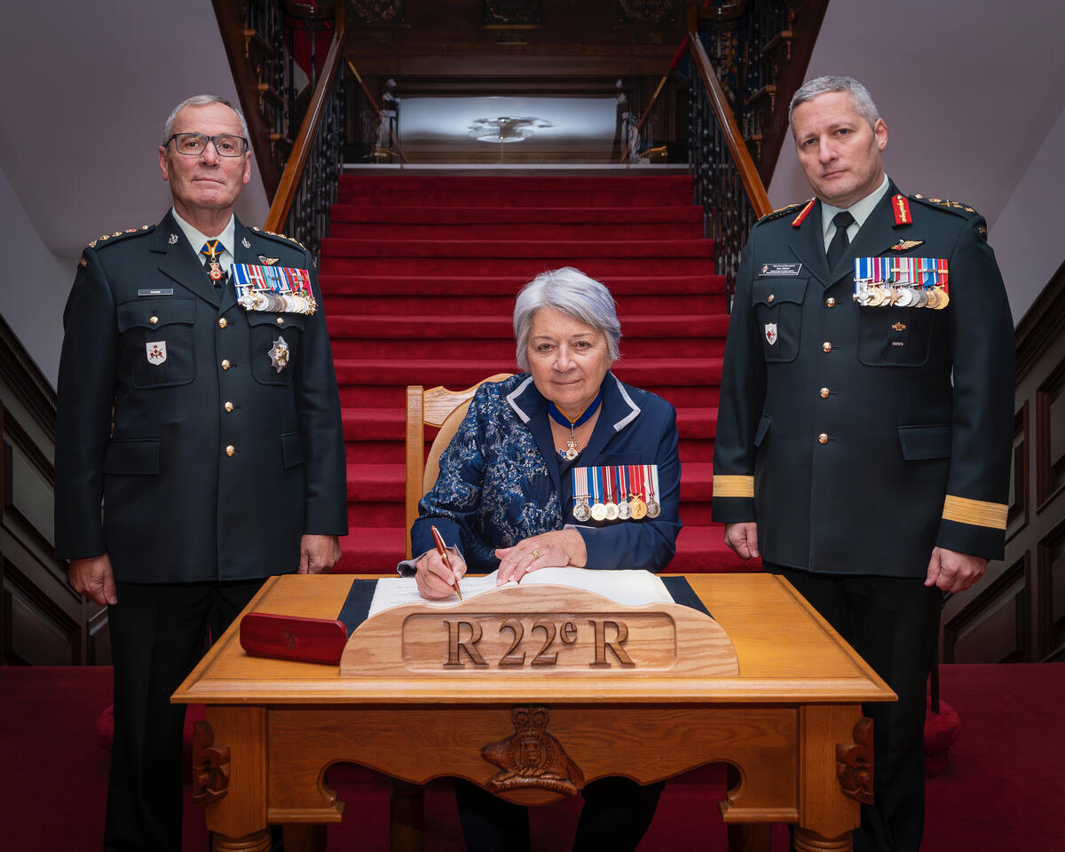 Governor General Mary Simon sitting at a desk signing a document, flanked by two men in military uniforms with medals. The setting is formal with a red carpeted staircase.
