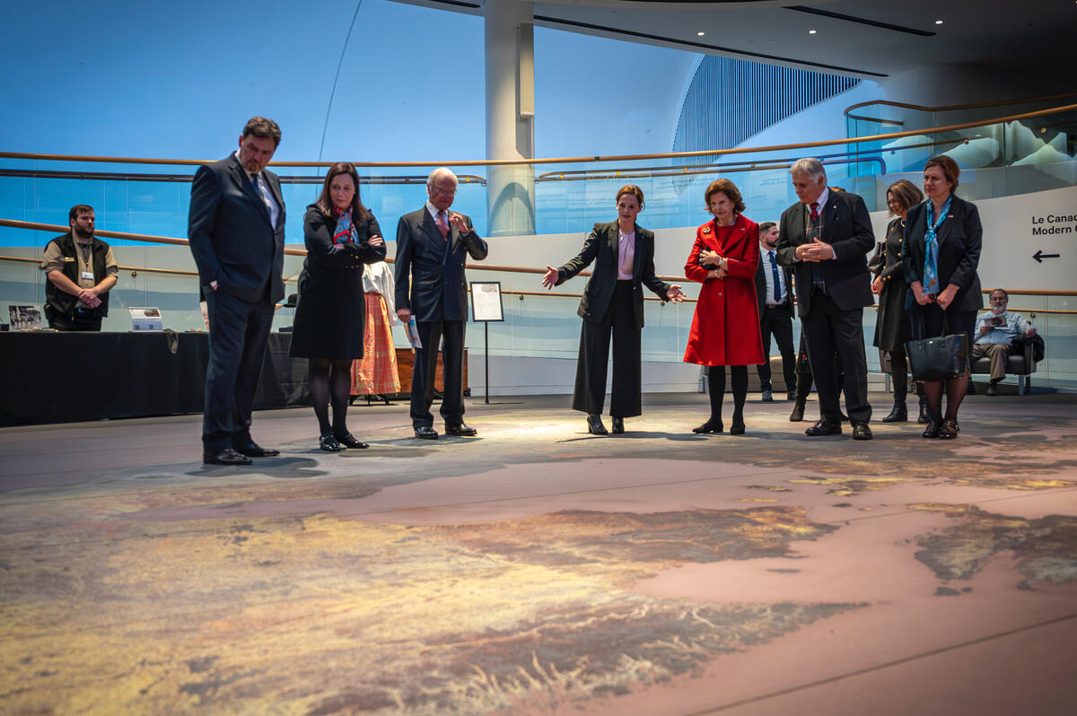 A museum employee is showing a large floor display to Their Majesties King Carl XVI Gustaf Queen Silvia of Sweden, the Right Honourable Richard Wagner, the Honourable Catherine Mandeville and Mr. Whit Fraser. 