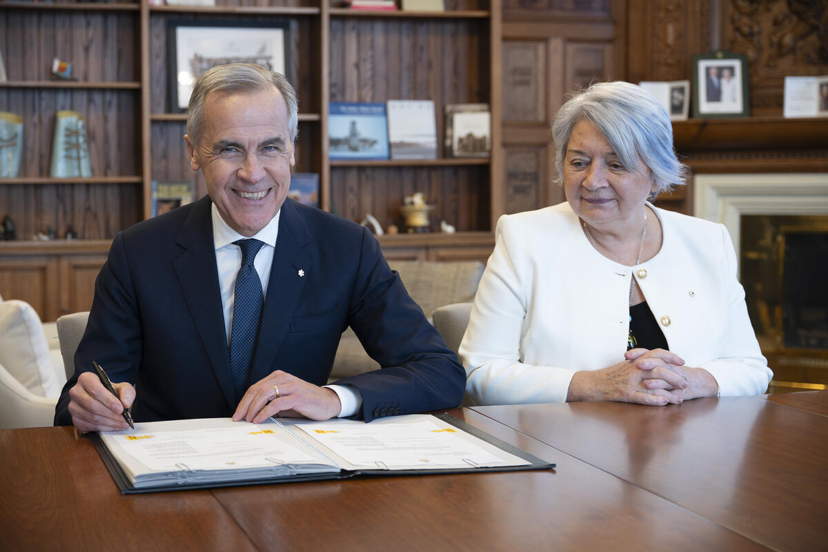 Le premier ministre Mark Carney et la gouverneure générale Mary Simon sont assis à une grande table en bois. Mark Carney signe un document.