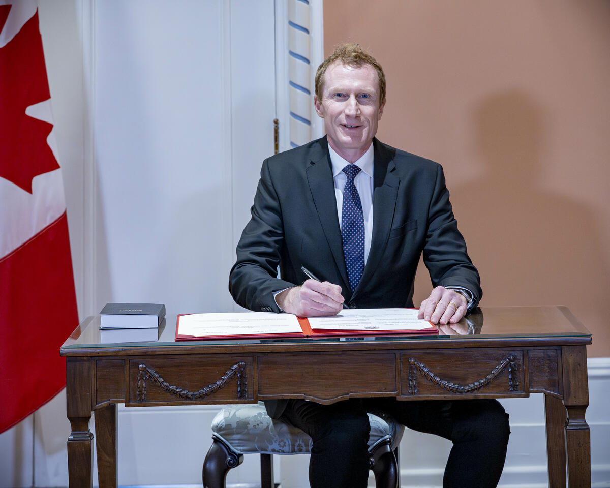 Mark Miller assis à un bureau en bois, en train de signer un document. À sa droite se trouve un drapeau canadien.