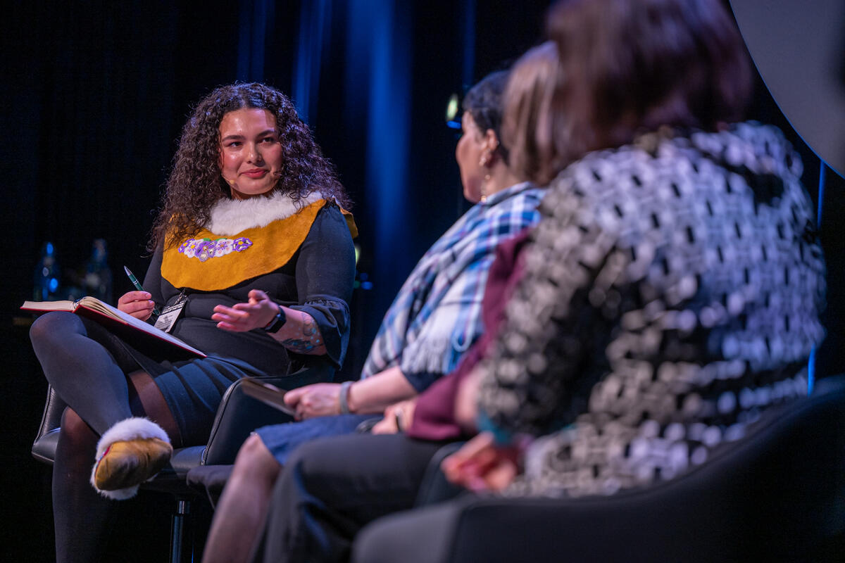 Une femme aux cheveux bouclés vêtue d'un costume traditionnel anime une table ronde avec trois femmes, toutes assises sur scène, devant un fond bleu faiblement éclair.