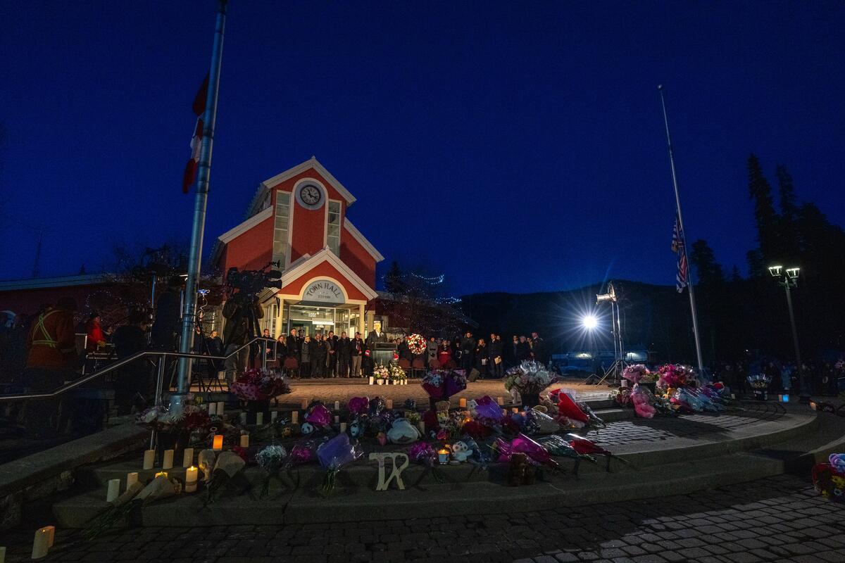 Le premier ministre du Canada, Mark Carney, prend la parole devant l’hôtel de ville de Tumbler Ridge, en Colombie-Britannique. Un groupe de personnes se tient derrière lui, et des fleurs et des bougies sont dispersées sur le sol.