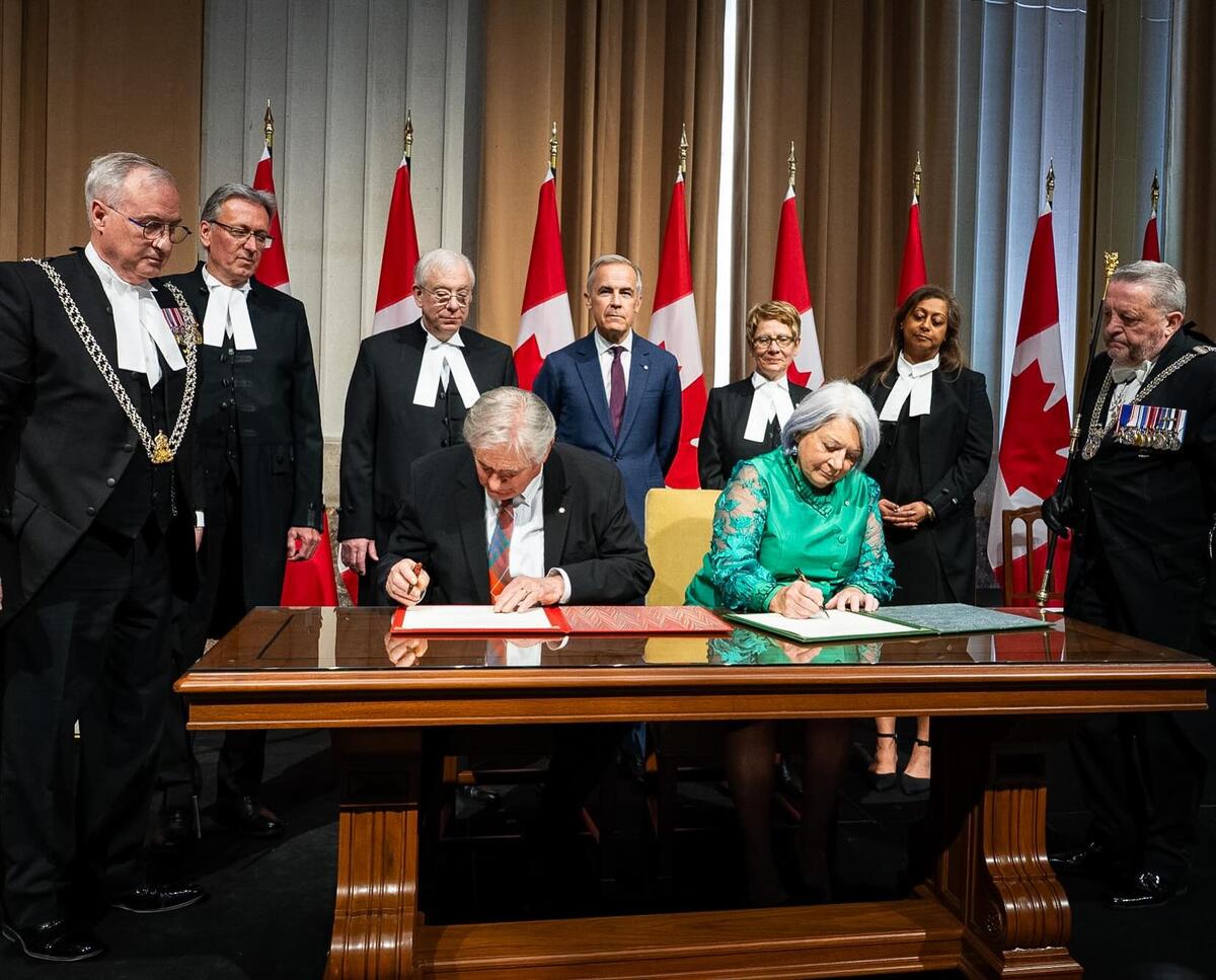 Governor General Mary Simon and Mr. Whit Fraser signing documents at a large table while people look on, including Prime Minister Mark Carney.