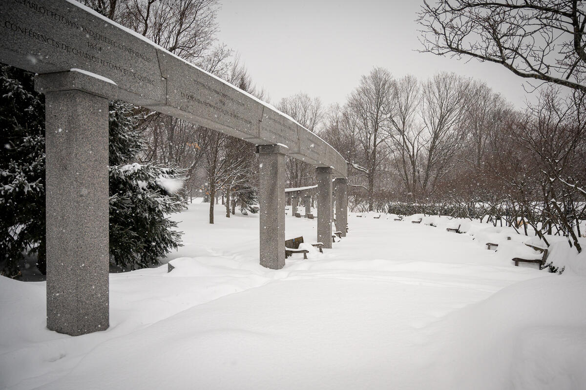 Snow-covered park with a long stone colonnade, bare trees, and benches blanketed in snow. The scene conveys a serene and tranquil winter atmosphere.